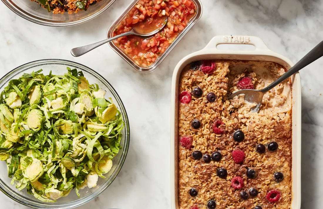 An overhead shot of a full week's healthy vegetarian meal prep, featuring diverse dishes like a black bean quinoa salad, fresh chopped salads, and a berry baked oatmeal.