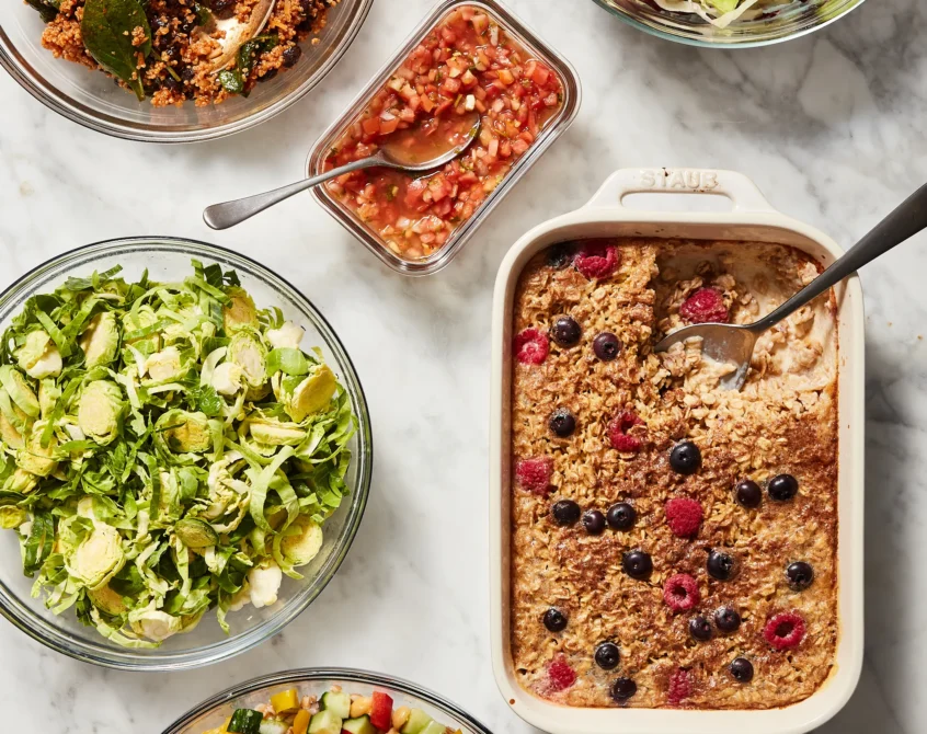 An overhead shot of a full week's healthy vegetarian meal prep, featuring diverse dishes like a black bean quinoa salad, fresh chopped salads, and a berry baked oatmeal.